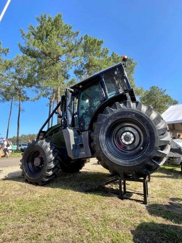 Tracteur forestier robuste noir surélevé sur un support, gros pneus, cabine sécurisée, sous un ciel bleu avec des pins en arrière-plan.